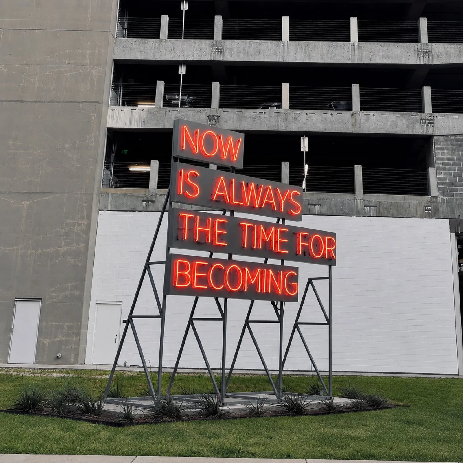 A red neon sign on angled steel frames reading "Now is always the time for becoming," in front of a concrete parking garage.
