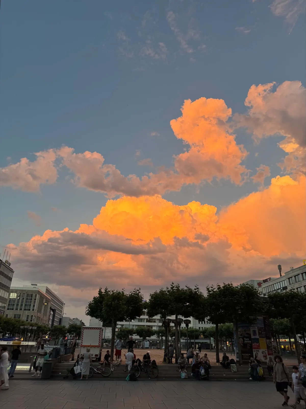 A city square at dusk. People sit and stand on the steps and pavement beneath a sky filled with towering cumulus clouds, their undersides lit orange by the setting sun.